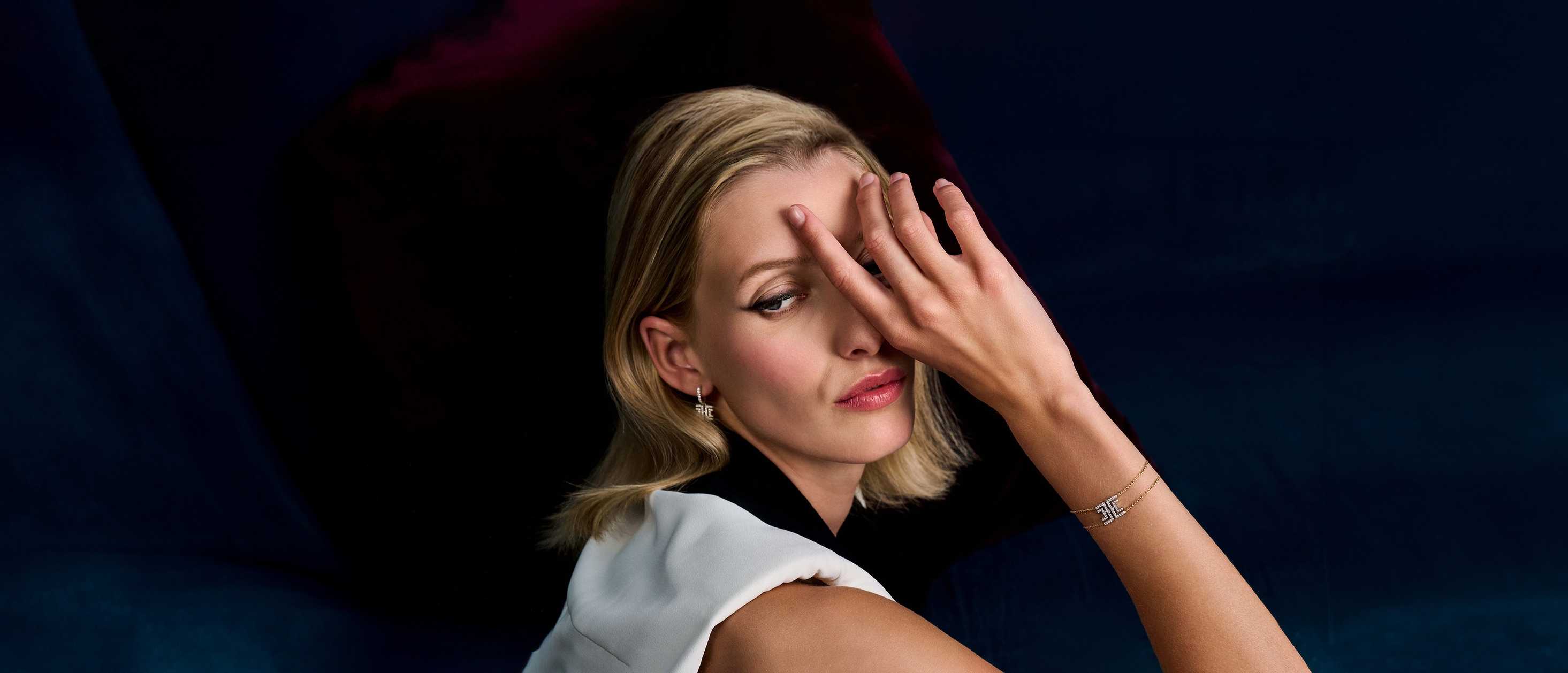 A model wearing the Harper Bracelet and matching earrings, featuring a geometric castle-set diamond centrepiece on a delicate double-chain in 18k gold. She poses with her hand partially covering her face, against a deep blue velvet background.