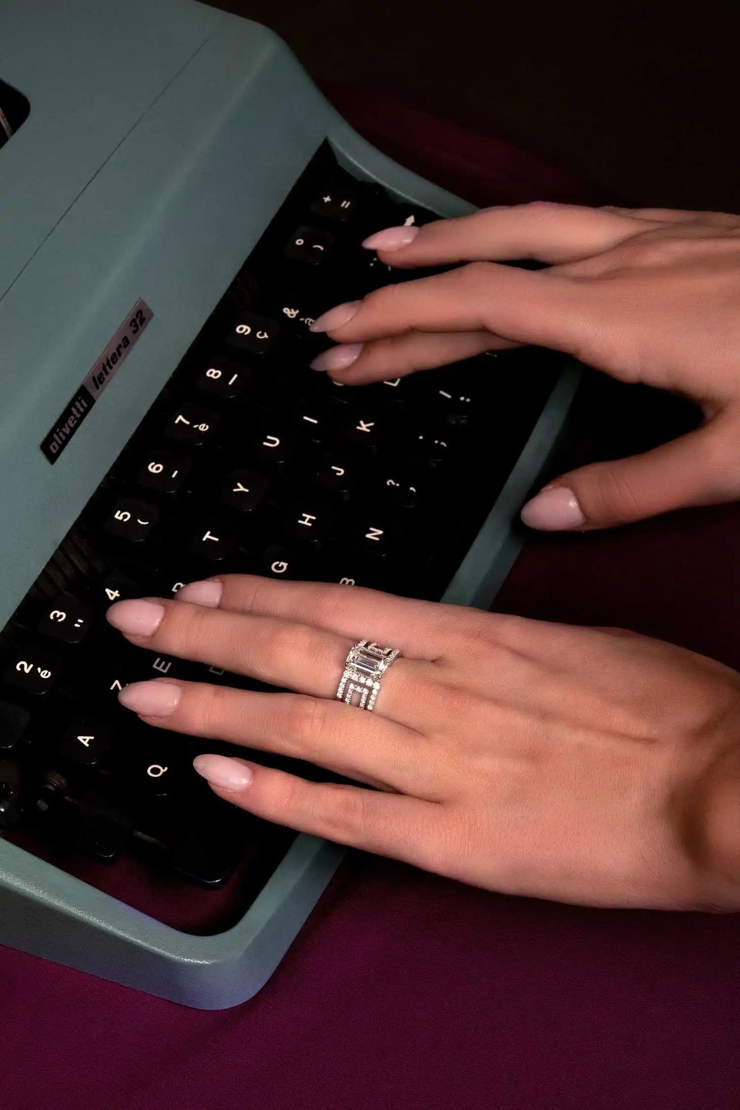 Hands typing on a vintage Olivetti Lettera 32 typewriter, adorned with the Heloise Emerald with its 3 ct Emerald cut.