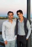 Brothers Ilan and Robin Okker standing together by the waterfront at sunset, dressed in light-striped shirts.