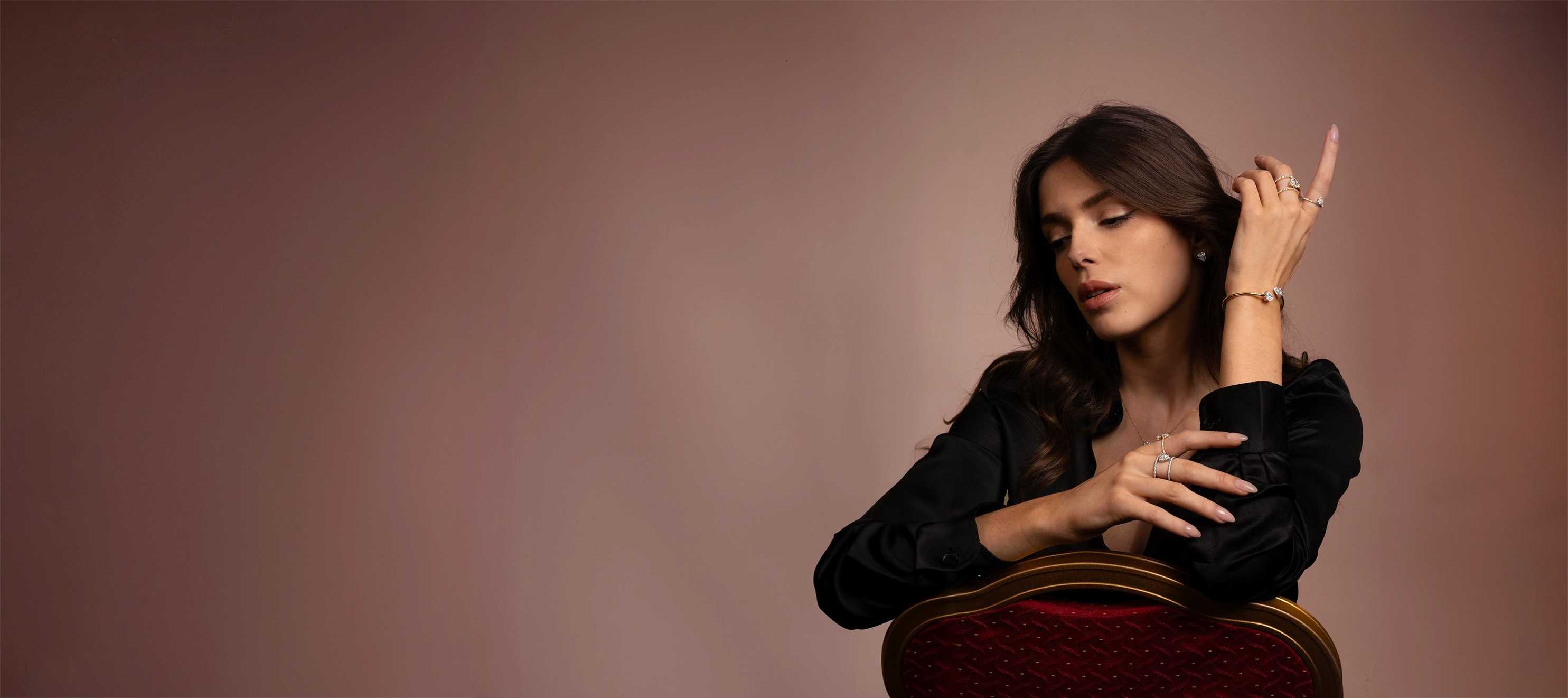 A model stacking Okker & Ockers lab-grown fine jewellery while posing with a chair, wearing rings, a bracelet, and studs.