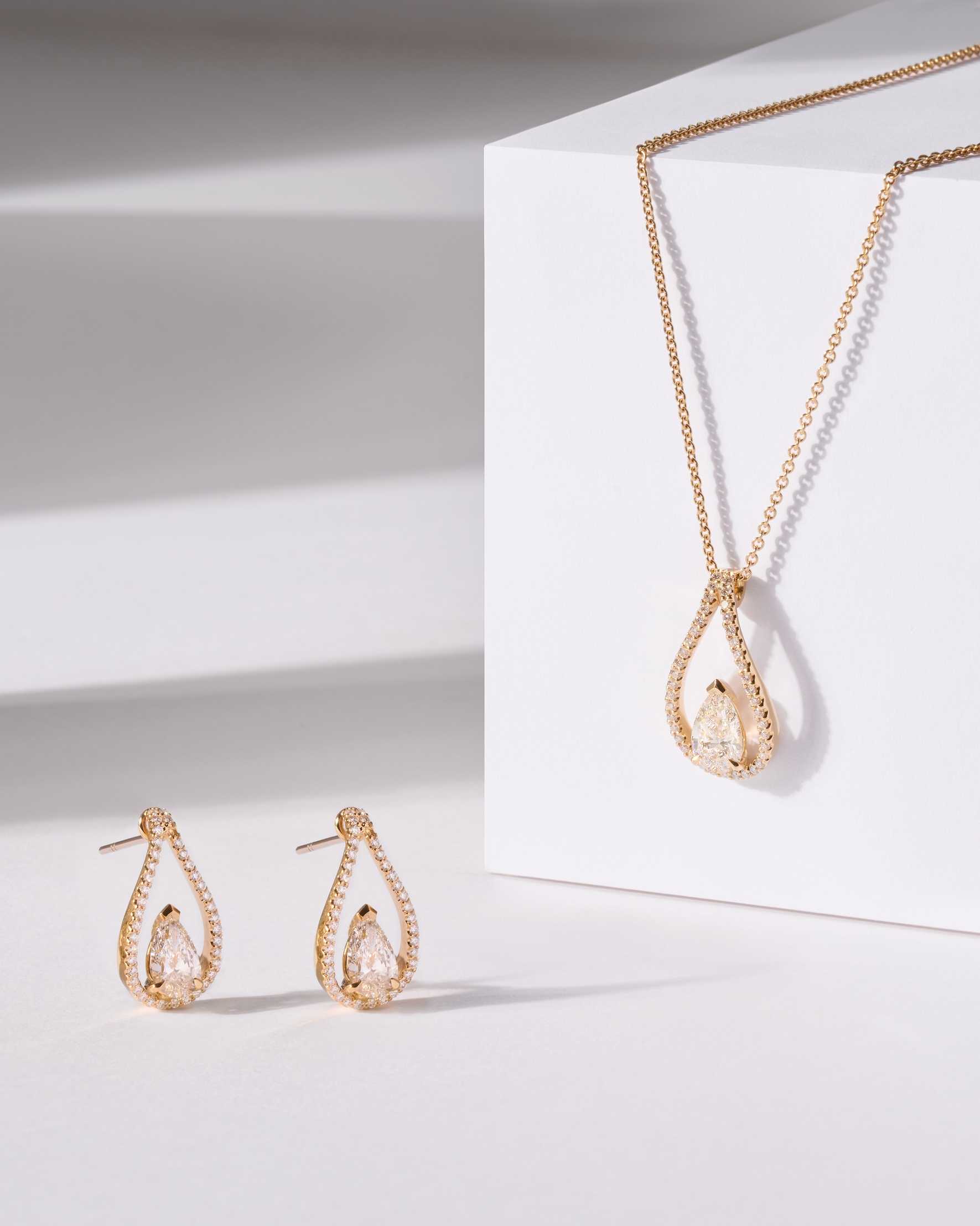 Floating Pear earrings and Floating Pear pendant in 18k gold displayed on a white and geometric background.